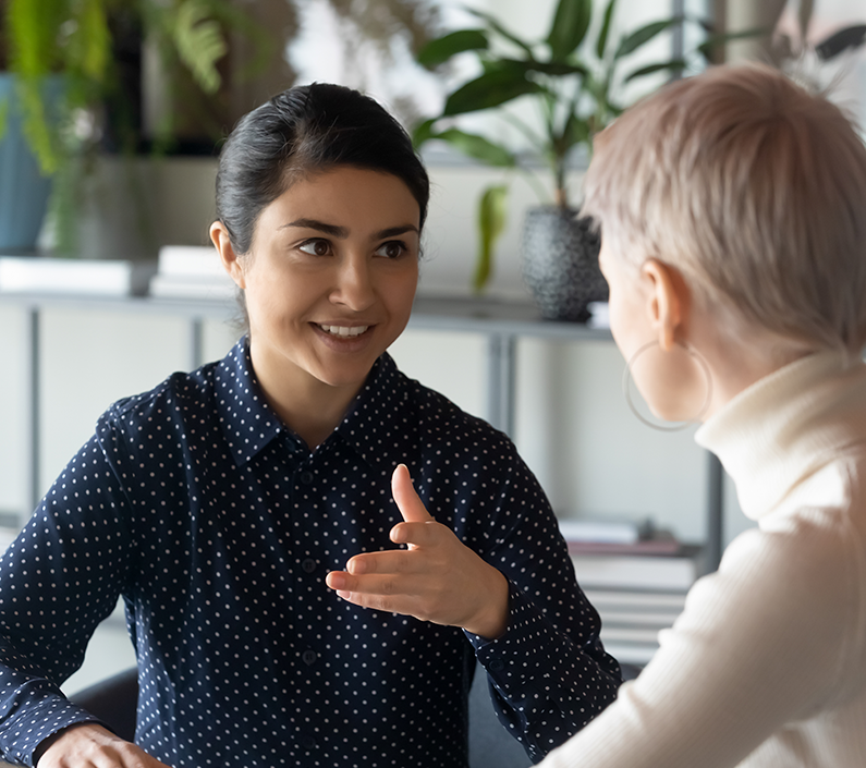 Friendly millennial businesswoman talking to blonde female colleague, sitting at table in office.
